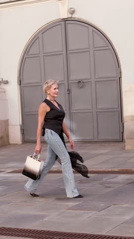 Elegant senior woman walking outdoors with stylish handbag, wearing sleeveless top and jeans, confident fashionable mature female lifestyle portrait in city street background