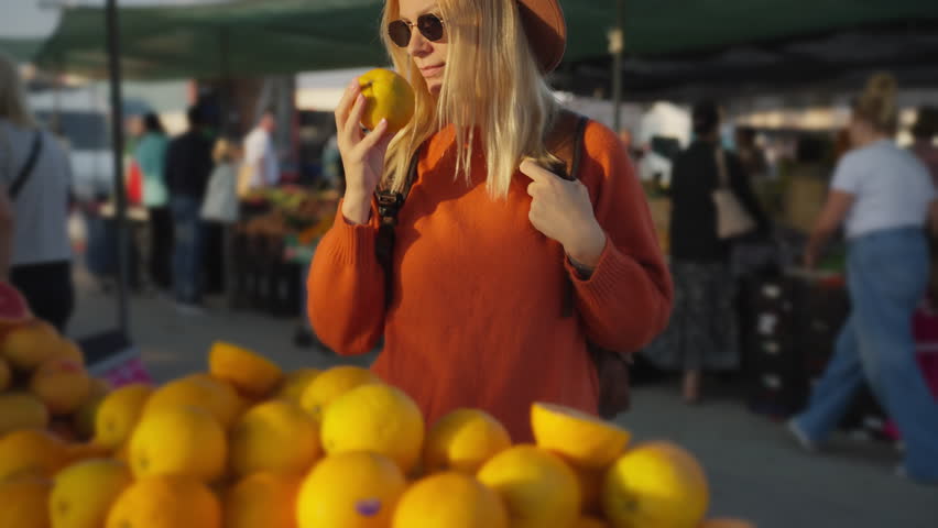 Beautiful young woman wearing sunglasses and beret smelling fresh lemon at busy outdoor market stall, enjoy aroma of citrus fruit before making purchase