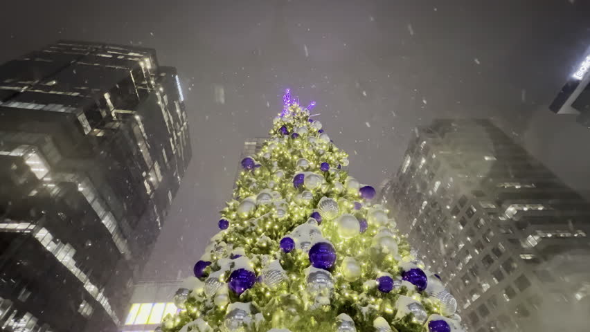 A towering Christmas tree, decorated with lights and ornaments, stands in a public plaza during a heavy winter snowstorm in downtown Rosslyn, VA. Snow falls softly against a backdrop of tall buildings