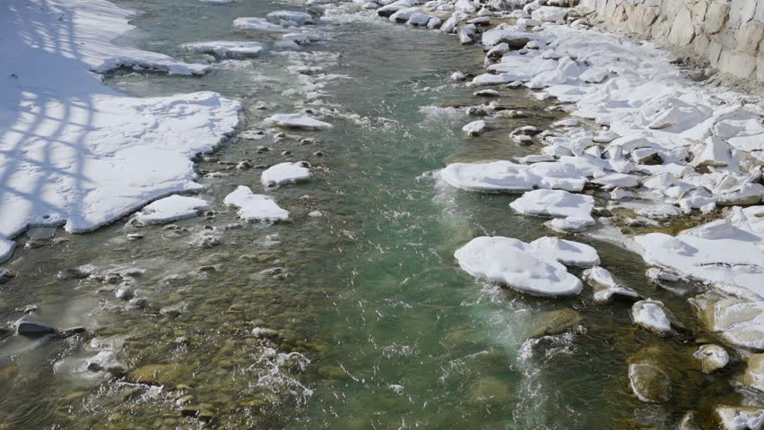Freezing cold water flowing down stream with pieces of white snow on winter day. Small town in European Alps with ski resorts and hotels. Countryside with fresh icy water straight from local mountains
