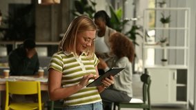 Young professional woman using a tablet, smiling while working in a busy, creative open plan office. Her multi ethnic team is collaborating in the background - Powered by Shutterstock - Get 15% off with code: PIKWIZARD15