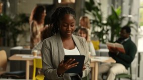 Young african american female professional using a digital tablet in a busy, modern open plan office, then smiling confidently at the camera - Powered by Shutterstock - Get 15% off with code: PIKWIZARD15