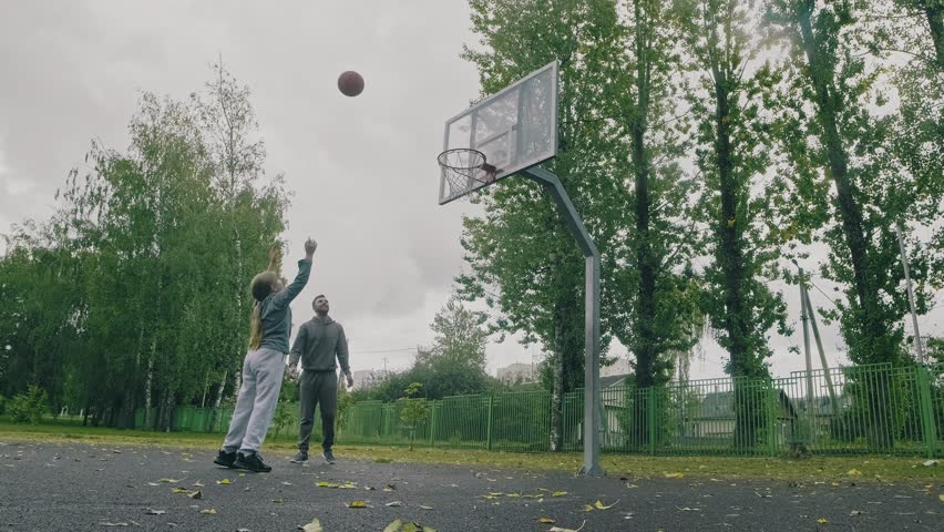 Cheerful child practices playing basketball outdoors. Young sportive father playing basketball with daughter. 4K, UHD
