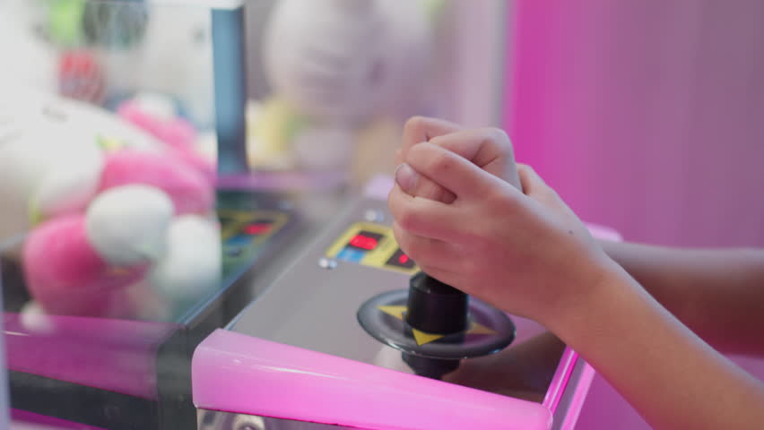Closeup of girl hands holding joystick while playing claw machine at arcade game center with stuffed toys inside creating fun playful atmosphere