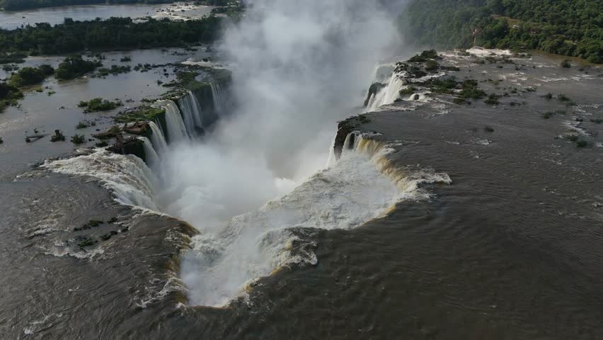 Aerial view soars above Iguazu Falls in Argentina. A vast river flows over the edge and dives into a giant canyon. White mist rises and fills the gorge with energy and natural beauty
