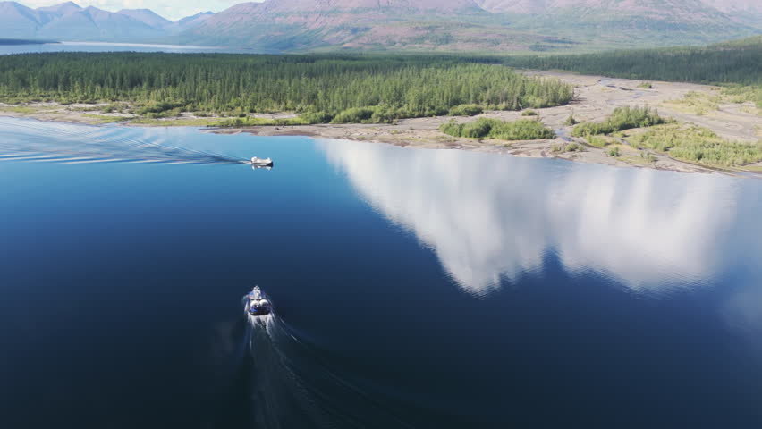 Glass water mirrors cloud as a motor boat crosses Lake Lama toward the delta. Mountains of the Putorana Plateau rise in blue distance. Silence, reflection and movement make a perfect travel scene