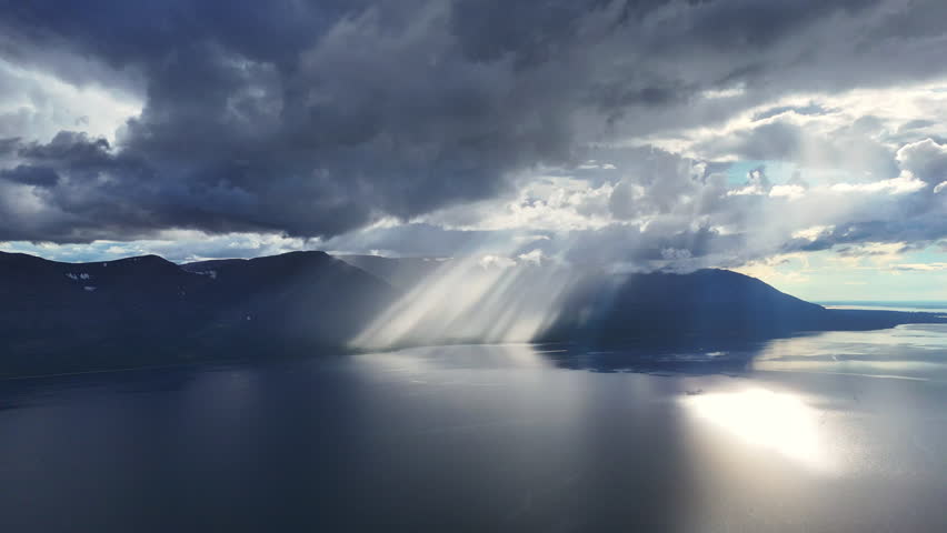 Rain core and slant light meet over the mountain wall by Lake Lama, forming bright veil on the water. Mist curls along the ridge, highlighting the raw Siberia landscape and timeless volcanic plateau