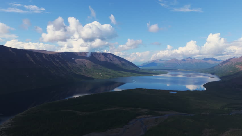 Aerial view of Lake Lama basin where ridge shadow slides across deep blue water. Bright cloud float above a green shore and distant peak. The panorama promises solitude, purity and adventure in Arctic
