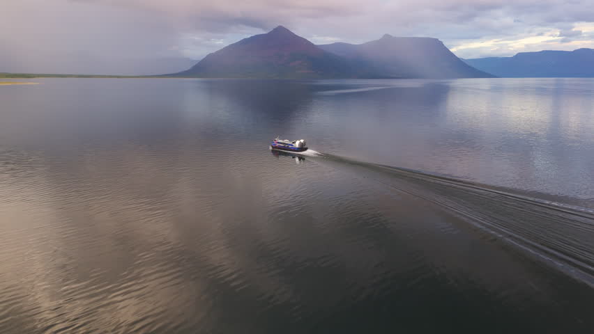 Remote boat glides across mirror water of Lake Lama as rain streaks soften slopes. Evening haze and long wake add drama and scale. Wilderness logistics and adventure travel in the Putorana Plateau