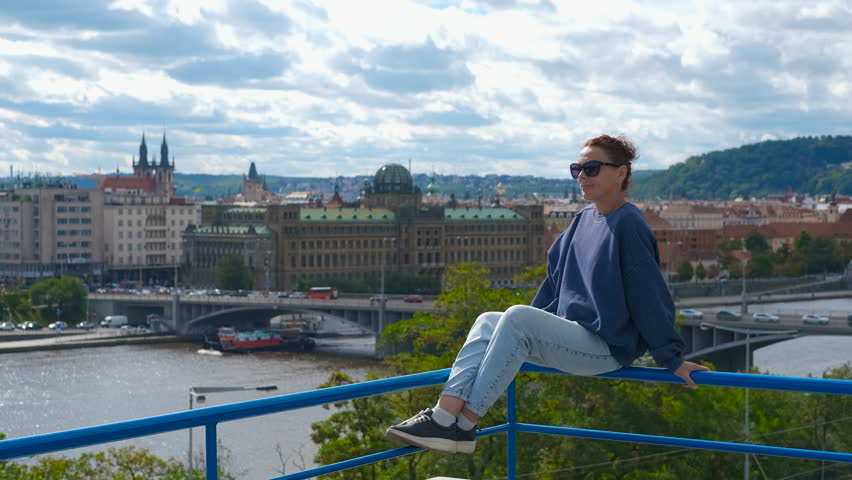 A woman looks at the cityscape of Prague. Young woman sitting on blue railing, overlooking historic prague cityscape with vltava river, iconic bridges, and architectural landmarks under sunny sky