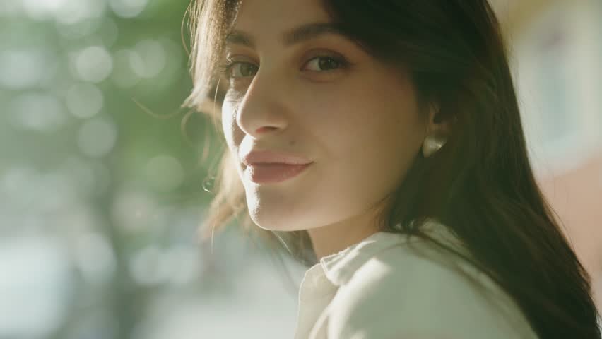 Close-up portrait of a woman with long hair, smiling warmly in a natural outdoor setting with soft sunlight filtering through trees in the background. Close-Up Portrait Of A Woman With Long Hair