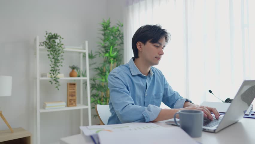 Asian attractive man freelancer using laptop device working in house. Handsome male student chatting and typing on computer notebook to communicate online with technology in living room at home.