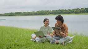 Asian young couple enjoying leisure time together outdoors in the park. Attractive man and woman sitting on the grass near lake, using a laptop and digital tablet, sharing ideas and relaxing in garden - Powered by Shutterstock - Get 15% off with code: PIKWIZARD15