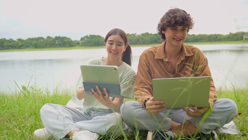 Asian young couple enjoying leisure time together outdoors in the park. Attractive man and woman sitting on the grass near lake, using a laptop and digital tablet, sharing ideas and relaxing in garden