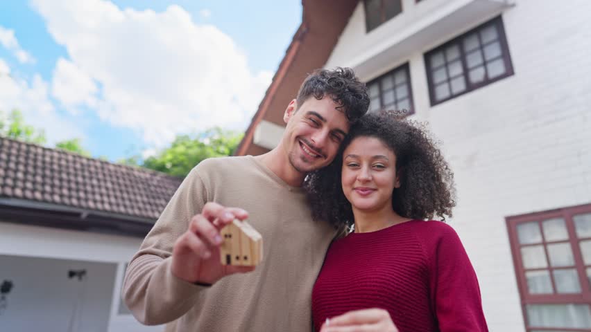 Hispanic Latina young couple holding a key and small wooden house model. Portrait of attractive man and woman standing outdoors infront of new home, symbolizing moving in, or real estate ownership.