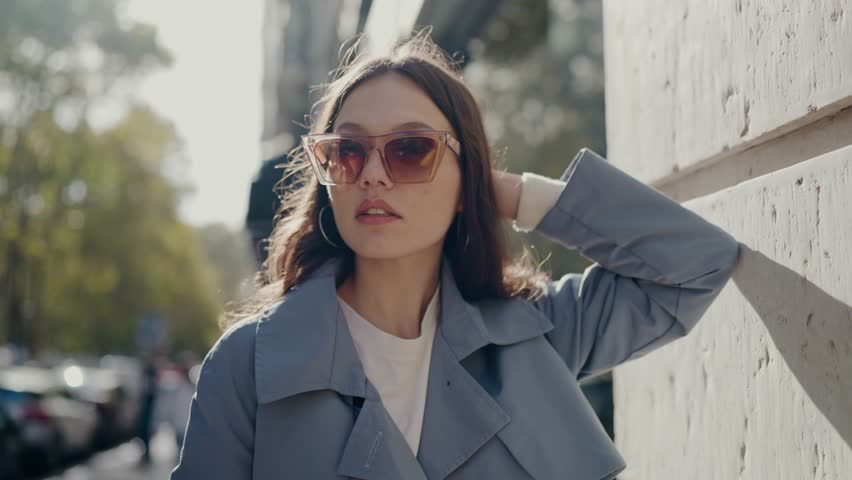 Stylish young woman with sunglasses walking confidently on a busy city street, touching her hair and enjoying the sunlight. Fashionable Young Woman in Sunglasses Enjoying Sunny City Day