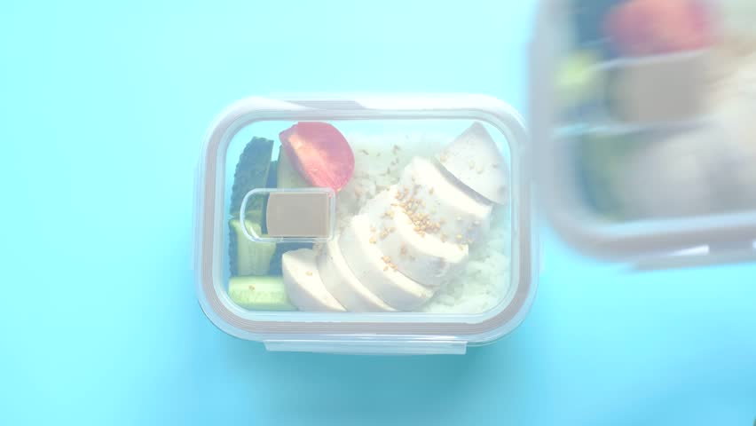A hand holds a glass box with rice, chicken and vegetables on a blue background, top view, close-up.