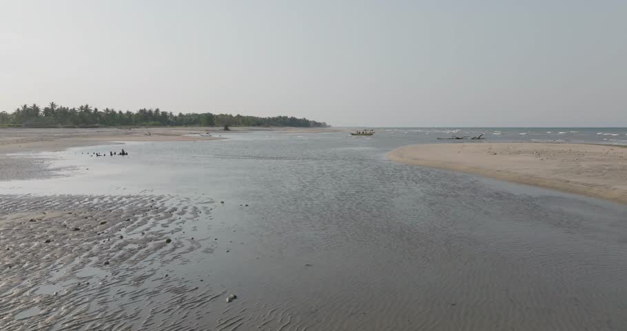 Low altitude drone flight over a serene tropical estuary where a river meets the ocean, revealing a vast sandy beach and a local boat on a calm, hazy morning in Southeast Asia
