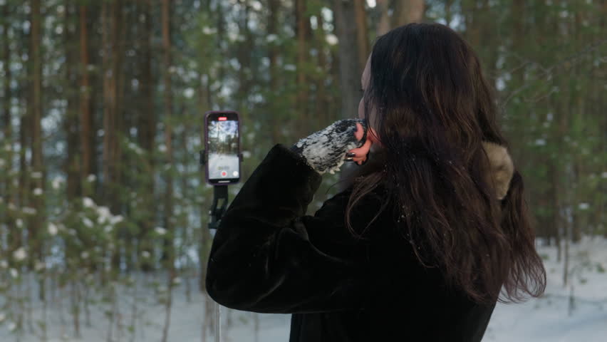 Woman in black coat and brown fur collar using smartphone on gimbal or selfie stick, filming video in snowy forest. Content creation, vlogging, back view.