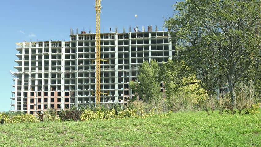 Large concrete residential building under construction with a tall yellow tower crane operating against a clear blue sky, seen from a green, grassy hill with lush trees on a sunny day