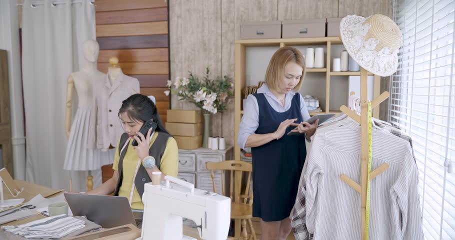 Asian female fashion designer discussing clothing detail with assistant while another young woman working on laptop near sewing machine in modern tailoring studio for small fashion business
