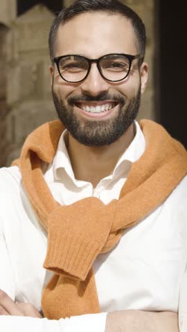 An Arab man smiles confidently outdoors, dressed in a white shirt and orange sweater, showcasing natural charm.