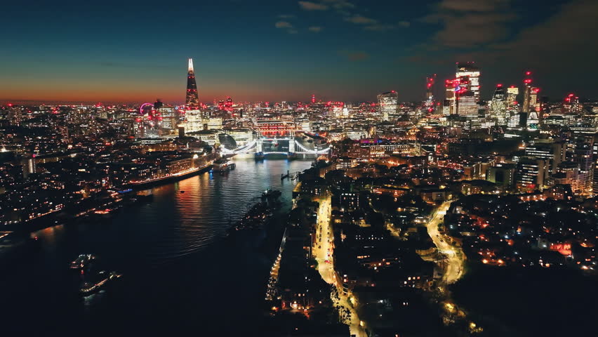 England, London: Breathtaking aerial view of night London city with Tower Bridge, The Shard, and other landmarks in colorful bright lights creating a stunning cityscape skyline under sunset sky