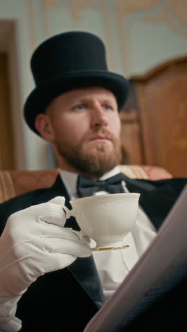 Vertical waist up shot of distinguished Caucasian nobleman in black tuxedo, cylinder hat, and white gloves sipping tea from white china cup while reading magazine in refined, classic setting