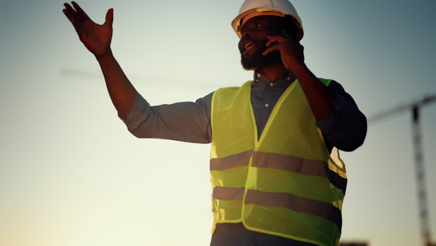 Boss discussing business by phone, architect or foreman on construction site. Portrait of african american male person with helmet and green vest against architectural engineering, cinematic shot