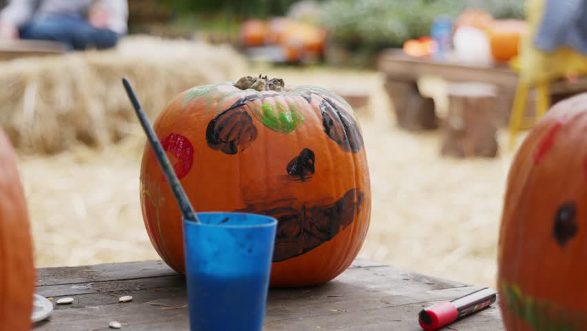 Close-up of a painted orange pumpkin decoration with a spooky black face, brush and paint cup on a wooden table for a fun Halloween activity