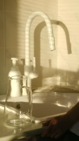 Cinematic shot person hands filling glass pitcher with clean drinking water from a stylish white tap in a sunny kitchen. Person filling glass pitcher with water from a modern kitchen faucet
