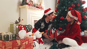 Mother and child in Santa hats open a glowing gift beside a decorated Christmas tree and fireplace, surrounded by wrapped presents, snowman toys, and cozy holiday decor. Real time 4k video - Powered by Shutterstock - Get 15% off with code: PIKWIZARD15