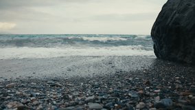 Low-angle shot of ocean wave approaching a rocky shore, with thick white foam covering the pebbled beach. A large dark cliff is visible on the right side. Overcast sky and horizon in the distance. - Powered by Shutterstock - Get 15% off with code: PIKWIZARD15