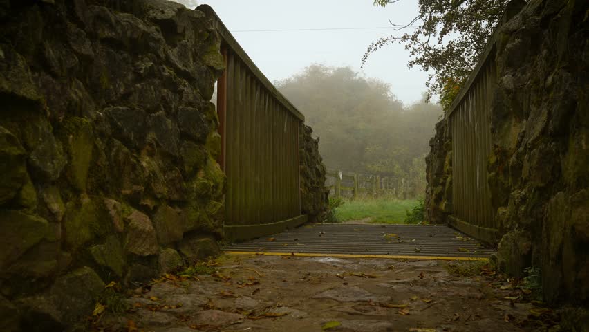 Old wooden bridge between mossy stone walls leading into misty countryside path. Morning fog merging with silence creating spiritual depth. Rustic walkway crossing into fog filled meadow surrounded by