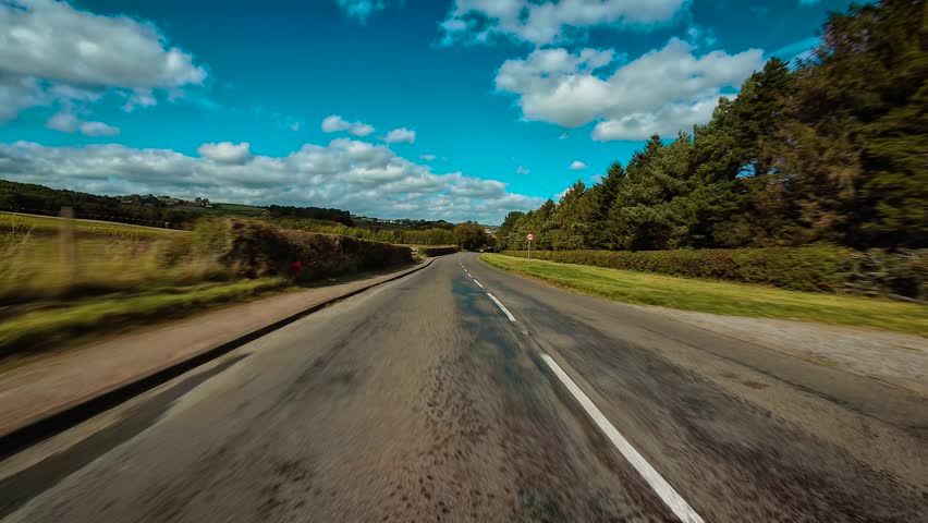 Driving along countryside road surrounded by green fields and trees under bright blue sky POV. Moving through rural asphalt road bordered by hedges and forest first person view. Traveling down scenic