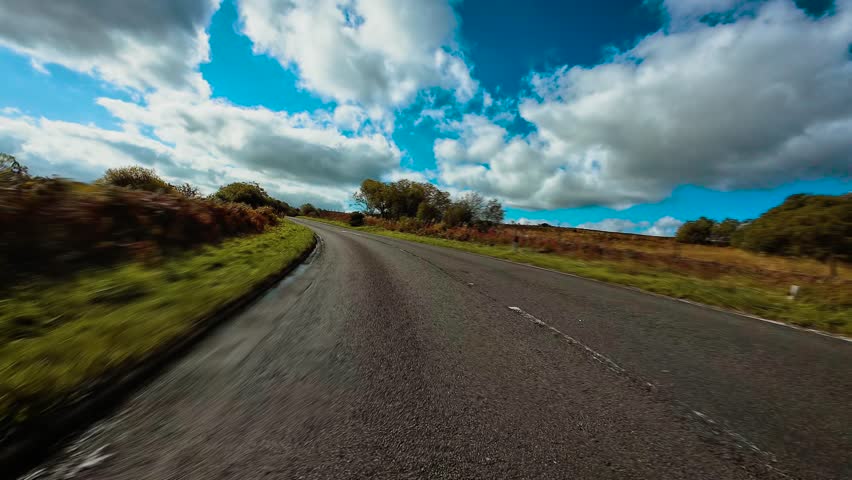 Car driving through countryside road with left hand traffic in England surrounded by open landscape POV. SUV moving along rural British highway bordered by grass and bushes under bright sky first