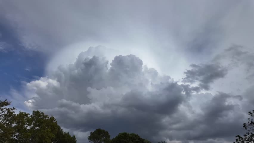 Timelapse of a threatening and massive storm cloud evolving in a turbulent blue sky over some trees. Daylight.