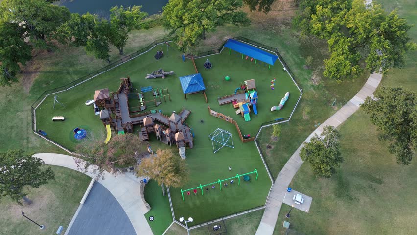 Playground in Highland Village featuring castle-style towers, colorful slides, swings, shaded pavilion. Enclosed by fencing, set on artificial turf with walkways, lakeside greenery, kids playing. USA
