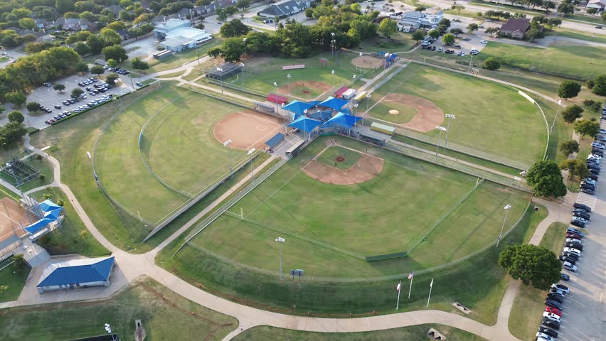 Fenced baseball fields active diamonds with active player, central pavilion, surrounded by paved trails, parking, clubhouses, tree-lined residential streets in Highland Village suburban corridor. USA