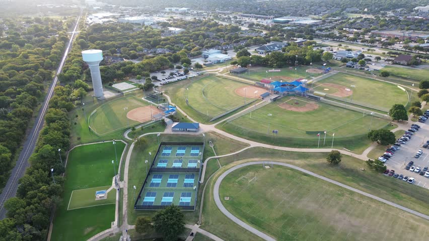 Expansive aerial of Highland Village sports complex with baseball diamonds, tennis pickle courts, soccer field, water tower, rail corridor, surrounded by trees, trails, and suburban zoning, Texas. USA