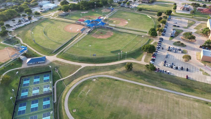 Full tennis pickleball courts with players, baseball softball fields cloverleaf with central blue-roofed pavilion, bleachers, paved paths. Adjacent to parking, trees, light commercial buildings. USA