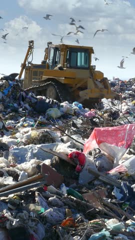 Garbage dump environmental pollution, earthmover bulldozer is working through massive piles of trash while many seagulls fly above. There are lots of plastic bags and bottles at landfill near the city