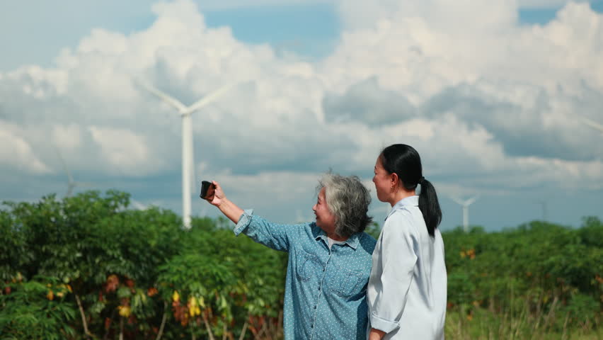 Two Asian female taking photo by smartphone. wind turbines in the background Representing Renewable Energy Eco Lifestyle and Sustainable Future. - Powered by Shutterstock - Get 15% off with code: PIKWIZARD15
