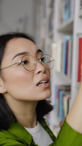 Asian woman looks through titles of books on library bookshelf carefully. Young teacher happy to find interesting book to read. Pursuit of knowledge closeup
