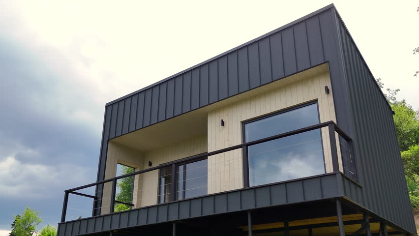 Modern cubic minimalist house exterior with dark metal siding and large glass windows overlooking a natural landscape