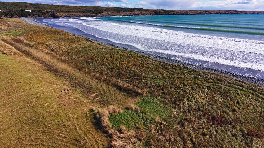 A sweeping drone perspective over Biscay Bay reveals rolling surf meeting a coastal meadow and grassy fields, with a winding shoreline and blue ocean stretching to the horizon.
