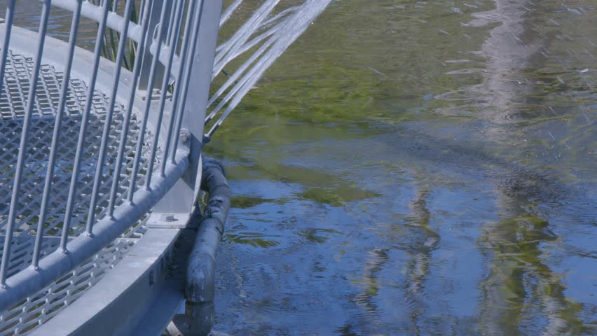 Serene close-up of a metal bridge railing with flowing water beneath, capturing reflections and gentle ripples. Sunlight enhances the peaceful, natural setting.