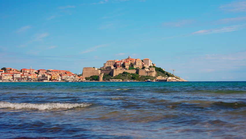 Static shot of the historic town of Calvi in Corsica, France. View of the bastion Citadel overlooking the town. Turquoise sea, waves coming to the shore. Summer holidays destination.