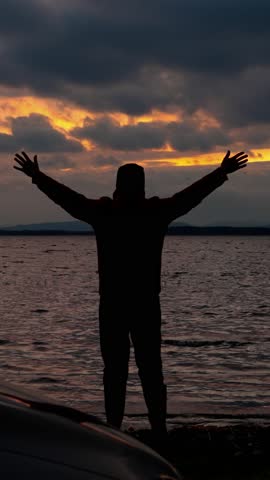 The silhouette of a man, admiringly raising his hands up and admiring the beautiful sunset on the shore of the lake. Symbolizing human happiness from natural beauty. Vertical video, shorts.