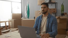 Young hispanic man in casual clothes works on laptop surrounded by moving boxes in new home living room setting. - Powered by Shutterstock - Get 15% off with code: PIKWIZARD15